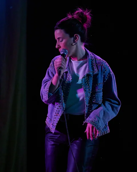 Micaela Lavin singing into a microphone under stage lighting.